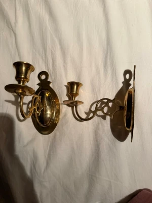 Guldiga väggljusstakar i mässing - Säljer ett par klassiska väggljusstakar i blank mässing. De har en oval bakplatta och snyggt svängda armar med dekorativa detaljer. Perfekta för att skapa mysig stämning och vintagekänsla i rummet. Passar vanliga kronljus och är enkla att hänga upp.