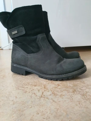 Svarta fodrade boots från Tamaris - Säljer ett par svarta boots från Tamaris med grov sula och dragkedja på sidan. Utsidan är i mocka och skinn, insidan är fodrad med mjuk pälsimitation. Snygga kontrastsömmar och en enkel remdetalj upptill. Perfekta för kalla dagar!