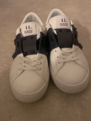 Valentino Rockstud vita sneakers - Säljer ett par vita Valentino Rockstud Untitled sneakers i skinn med svarta nitar och bred svart rem över vristen. Klassisk rund tå, vita snören och coola detaljer längs sidorna och hälen. Perfekta för dig som vill sticka ut med en edgy touch.