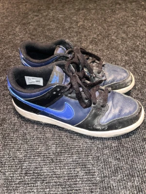 Nike Dunk sneakers blå och svart - Säljer ett par Nike Dunk sneakers i blått och svart med vit sula. Skorna har klassisk låg siluett, svart snörning och rund tå. Materialet är en mix av skinn och syntet. Perfekta för dig som gillar streetwear och vill ha en ikonisk sneaker i garderoben. Anledningen till det låga priset är pågrund av den lilla storleken! Tveka inte på att höra av dig om du har fler frågor!🤩🔥