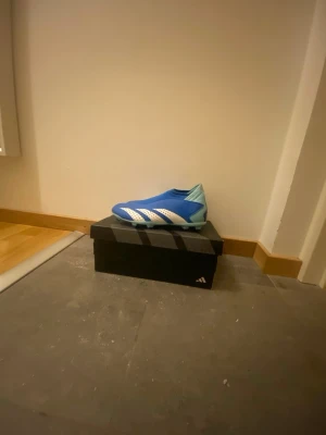 Säljer Adidas Predator blå fotbollsskor - Säljer ett par Adidas Predator fotbollsskor i blått med vita och röda detaljer. Skorna har en strömlinjeformad design utan snörning, ljusblå sula med dobbar och klassiska vita Adidas-ränder på sidan. Perfekt för dig som vill ha grym kontroll på planen.