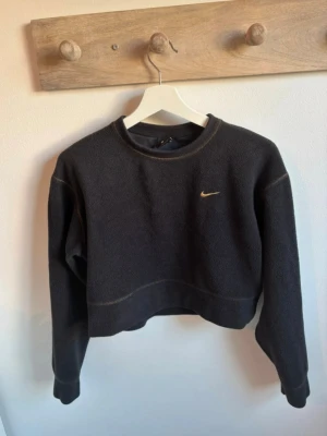 Svart fleece tröja från Nike - Mysig svart fleece tröja från Nike med guld broderad swoosh på bröstet. Tröjan har croppad passform, rund halsringning och synliga sömmar i kontrastfärg. Perfekt för kyliga dagar och sportiga outfits.