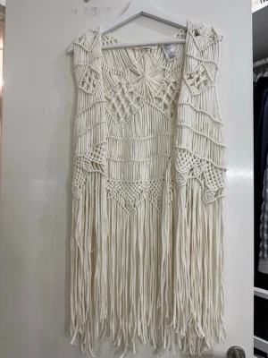 Vit boho väst med fransar - Säljer en vit väst i boho-stil med långa fransar och detaljerad knytning. Västen är gjord i tjockt bomullssnöre och har ett unikt mönster med olika geometriska former. Perfekt för dig som vill sticka ut med en cool och kreativ look.
