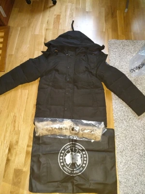 Svart dunjacka från Canada Goose - Svart dunjacka från Canada Goose med huva och avtagbar pälsdetalj. Jackan har långa ärmar, knappar med logotyp och klassisk patch på ärmen. Perfekt för kalla dagar och har en stilren, rak passform.