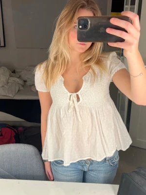 Vit blus med knyt och puffärm - Supersöt vit blus med korta puffärmar och knyt framtill. Blusen har smock över bysten och ett lätt, flowy tyg med diskreta prickar. Perfekt till jeans eller kjol för en fräsch look.