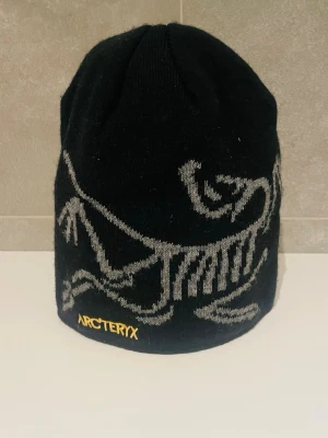 Arc’teryx beanie  - Säljer en svart mössa från Arc'teryx, modell Fernie Toque. Mössan har ett stort grått skelettmönster och gul Arc'teryx-logga broderad framtill. Tillverkad i mjukt material som passar perfekt för kalla dagar.