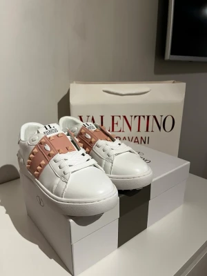 Valentino sneakers - Säljer ett par Valentino sneakers i vitt skinn med ljusrosa band och ikoniska nitar. Skorna har rund tå, snörning och platt sula. Perfekta för dig som vill ha en lyxig och edgy touch till din outfit.