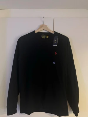 Svart tröja från Polo Ralph Lauren - Klassisk svart långärmad tröja från Polo Ralph Lauren i bomull. Tröjan har rund halsringning och den ikoniska röda logotypen broderad på bröstet. Enkel och stilren design som passar till allt.