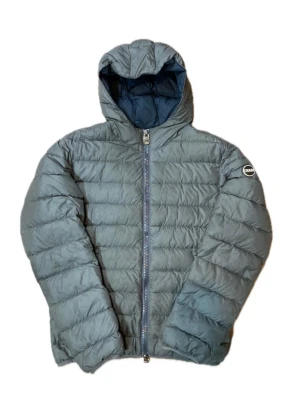 Grå pufferjacka från Colmar - Säljer en grå pufferjacka från Colmar med huva och hel dragkedja framtill. Jackan har ett quiltat mönster och en rund logotyp-patch på ärmen. Perfekt för kyliga dagar och riktigt skön att bära.