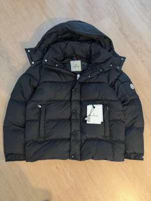 Svart Moncler pufferjacka med huva - Säljer en svart pufferjacka från Moncler med ikonisk logga på ärmen och stor huva. Jackan har dragkedja och knappar framtill, samt praktiska fickor. Insidan har ett coolt serietryck och jackan är fylld med dun för extra värme. Perfekt för kalla dagar.
