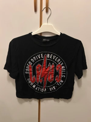 Svart croppad topp från PrettyLittleThing - Cool svart croppad topp från PrettyLittleThing med tryck i rött och vitt framtill. Rund hals och korta ärmar. Trycket har en blixtdetalj och texten 'Rodeo Drive Beverly Hills'. Perfekt för dig som gillar edgy streetstyle.