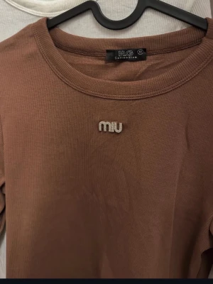 Brun ribbad långärmad topp MIU - Säljer en brun ribbad långärmad topp från MIU Collection. Toppen har rund halsringning och ett glittrigt 'miu'-emblem framtill. Materialet är mjukt och stretchigt, perfekt för en chill och snygg look.