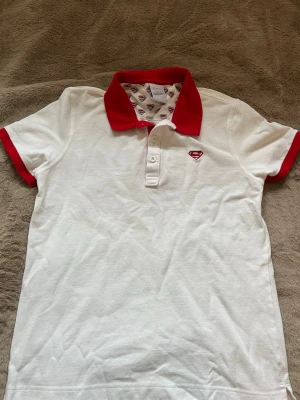 Superman vit och röd piké från Warner Bros - Vit pikétröja med röda detaljer från Warner Bros. Röd krage och ärmslut, broderad Superman-logga på bröstet och trycket 'LOOK! UP IN THE SKY!' på ryggen. Passar män och kvinnor, storlek M