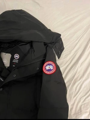 Svart dunjacka från Canada Goose - Säljer en svart dunjacka från Canada Goose med huva och tydlig logga på ärmen. Jackan har dragkedja och knappar framtill, samt flera praktiska fickor. Perfekt för kalla dagar och riktigt snygg till vinterlooken.