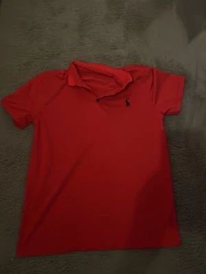 Röd piké från Ralph Lauren - Säljer en klassisk röd pikétröja från Ralph Lauren med mörkblå broderad logga på bröstet. Skjortan har korta ärmar, krage och knappar vid halsen. Perfekt till jeans eller shorts för en clean och sportig look.