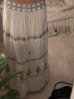 Vit långkjol från Zara - Supersnygg vit långkjol från Zara med svart broderat mönster och snörning i midjan. Kjolen har flera dekorativa band med blommiga detaljer och är gjord i ett lätt och luftigt bomullstyg. Perfekt för en chill och bohemisk vibe.