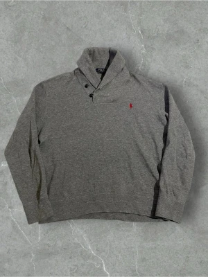 Grå tröja från Polo Ralph Lauren | M - Säljer en grå långärmad tröja från Polo Ralph Lauren med hög krage och knappar vid halsen. Tröjan har en liten röd logga broderad på bröstet och är tillverkad i mjuk bomull. Perfekt för kyliga dagar och enkel att matcha med jeans.