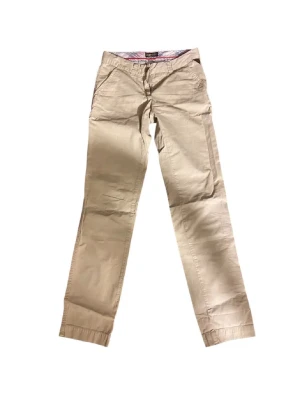 Beige chinos från Replay - Snygga beige chinos från Replay med raka ben och klassiska fickor fram och bak. Byxorna har bälteshällor och är tillverkade i mjuk bomull som känns skön mot huden. Perfekta för en clean och avslappnad stil.