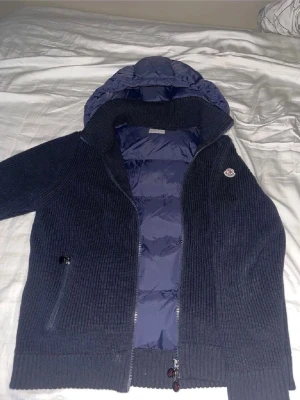 Mörkblå Moncler pufferjacka med huva - Säljer en mörkblå pufferjacka från Moncler med stickade ärmar och quiltad kropp. Jackan har en stor huva, dragkedja framtill och två fickor med dragkedja. Perfekt för kyliga dagar och riktigt snygg till jeans eller sweatpants.