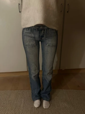 Blå bootcut jeans med stora fickor - Snygga blå jeans med bootcut passform och stora fickor både fram och bak. Jeansen har slitna detaljer och en cool tvättad look. Materialet är klassisk jeans och midjan är låg. Perfekta för dig som gillar retrostil och vill sticka ut.