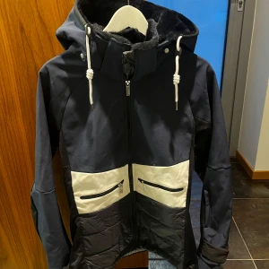 Svart och vit skidjacka från CMP - Snygg svart och vit skidjacka från CMP med huva fodrad i mjuk pälsimitation. Jackan har två framfickor med dragkedja och coola snörningsdetaljer vid huvan. Materialmix med polyester och syntet, perfekt för kyliga dagar.