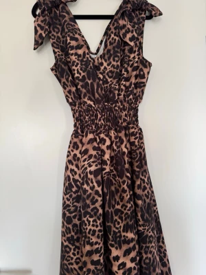 Leopardmönstrad omlottklänning - Säljer en snygg leopardmönstrad klänning med omlottdesign och bred resår i midjan. Klänningen har knytband på axlarna och är ärmlös. Tyget är mjukt och följsamt, perfekt för dig som vill sticka ut med ett djuriskt mönster.