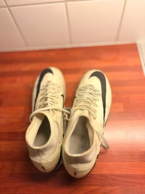 Nike Mercurial Air Zoom fotbollsskor - Säljer ett par vita Nike Mercurial Air Zoom fotbollsskor med svart swoosh och snörning. Skorna har högre strumpkrage för extra stöd runt ankeln och mönstrad ovandel i syntetmaterial. Sulan har fasta dobbar för bra grepp på planen. Perfekta för dig som vill ha speed och kontroll.