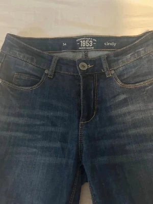 Blå bootcut jeans Cindy 1953 - Säljer ett par klassiska blå jeans från Cindy 1953 med bootcut passform. Jeansen har normal midja, snygga slitningar framtill. Tillverkade i mjukt denimtyg med kontrastsömmar för en tidlös look.