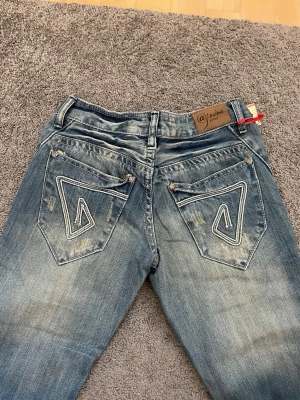 Blåa jeans med broderade bakfickor - Säljer ett par blå jeans med snyggt slitna detaljer och broderade bakfickor. Jeansen har klassisk femficksdesign, bootcut och låg midja. Materialet är jeans i bomull och de har en cool tvättad look. Säljer då de är lite små på mig, skriv vid intresse ❤️