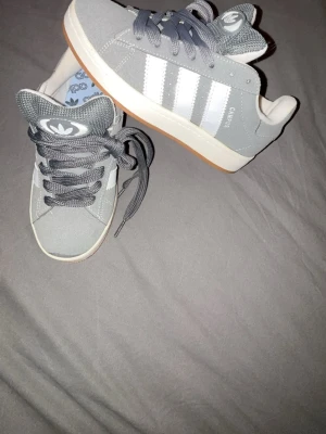 Grå Adidas Campus sneakers strl 38 - Säljer ett par grå Adidas Campus sneakers i storlek 38. Säljer dessa skor för att dom inte har kommit till användning har haft dom i över ett år, som sagt är dom aldrig använda så dom är i jätte bra skick. Skorna har klassiska vita ränder på sidan, grå snörning och en sula i gummi. Ovandelen är i mocka och insidan har Adidas-logga på innersulan. Perfekta för dig som gillar retrostil och streetwear. Dessa är då inte äkta då min pappa köpte dom när han var utomlands, PRISER GÅR ATT DISKUTERA! 