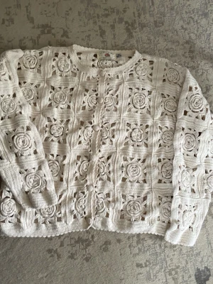 Virkat beige kofta med blommönster - Unik virkad kofta i beige med stora blommiga detaljer och genombrutet mönster. Koftan har lång ärm, rund hals och knappar framtill. Perfekt för dig som vill sticka ut med en bohemisk och kreativ stil.