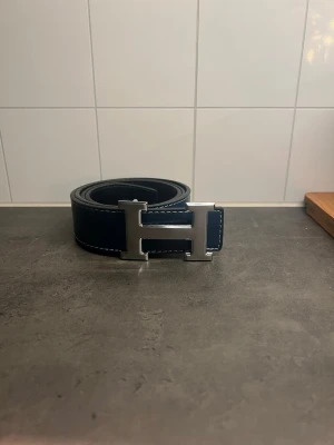 Herpés bälte - Bilderna visar ett blått bälte med ett stort H-format spänne i metall, men ingen väst finns med på bilderna.