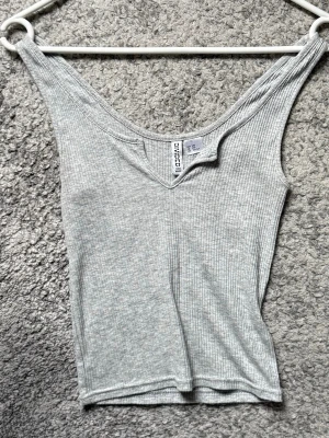 Ljusgrå ribbad linnetopp  - Säljer en ljusgrå ribbad linnetopp från Divided. Toppen har en v-ringad hals och är ärmlös med smala axelband. Materialet är mjukt och stretchigt, perfekt för varma dagar eller som lager under en skjorta.