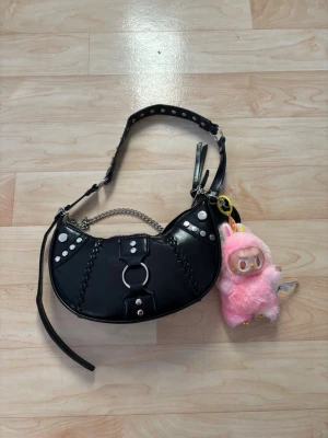 Svart axelväska med nitar och kedja - Säljer en svart axelväska i syntet med coola silverfärgade nitar, ringdetalj och kedja. Väskan har justerbart axelband med hål och dragkedja upptill. Medföljer en gullig rosa plysch-nyckelring. Perfekt för dig som gillar edgy detaljer.