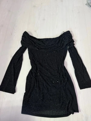 Svart glittrig offshoulder klänning - Säljer en svart kortklänning med glittriga detaljer och offshoulder-modell. Klänningen har långa utsvängda ärmar och sitter tight mot kroppen. Perfekt till utekvällar! Storlek L men skulle mer säga M! 