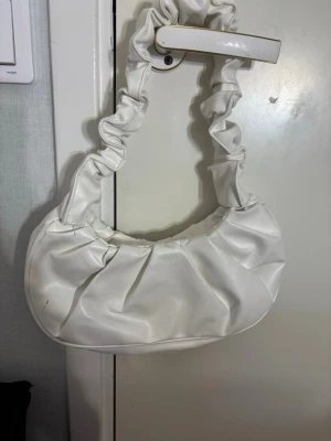 Vit axelväska med scrunchie-band - Säljer en superfin vit axelväska med scrunchie-inspirerat band. Väskan har en mjuk, rundad form och är gjord i syntetmaterial som ger en clean och modern look. Perfekt för dig som vill ha en trendig accessoar till din outfit.