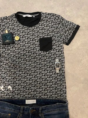 Svart t-shirt med bladmönster  - Svart t-shirt med vitt bladmönster över hela tröjan och svart bröstficka. T-shirten har rund halsringning och korta ärmar med svarta muddar. Tillverkad i mjuk bomull, perfekt för en chill och stilren look. Endast tröjan tillkommer! pris går att diskutera litegrann😃 S/XS. Tröjan har inget tillkännande märke, köptes utomlands