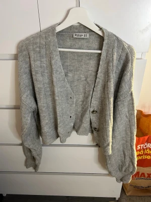 Grå ribbad kofta från Stradivarius - Mysig grå kofta från Stradivarius i ribbad stickad kvalitet. Modellen är croppad med v-ringning och stora knappar framtill. Perfekt till lager på lager och har långa ärmar med breda muddar. Enkel att styla till olika outfits. Priset kan diskuteras 🫶