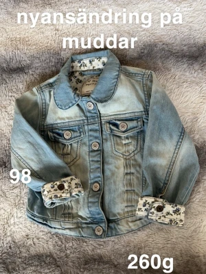 Ljusblå jeansjacka med blommiga muddar - Säljer en ljusblå jeansjacka från Next med snygga blommiga muddar och dekorativa knappar. Jackan har klassisk krage, två bröstfickor och långa ärmar. Perfekt för dig som vill ha en unik touch på din outfit!🌸 Liten nyansändring på muddarna. Storlek 98 