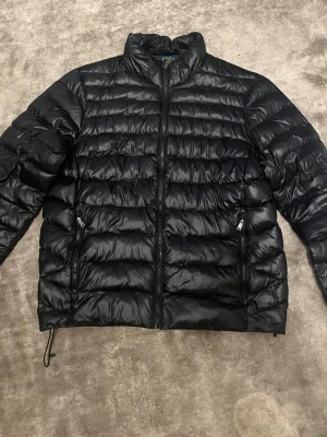 Svart pufferjacka från Polo Ralph Lauren - Snygg svart pufferjacka från Polo Ralph Lauren med hög krage och två sidofickor med dragkedja. Jackan har quiltad design och är tillverkad i glansig polyester. Perfekt för kalla dagar och har en clean look som funkar till många stilar.