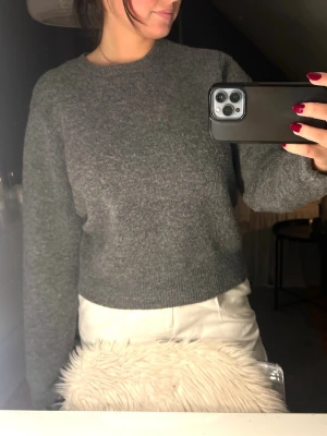 Grå stickad tröja med rund hals - Mysig grå stickad tröja med rund halsringning och lång ärm. Tröjan har en enkel och klassisk design som passar perfekt till jeans eller kjol. Den är mjuk och skön, perfekt för kyliga dagar.