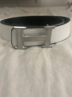 Svart och vit skärp med H-spänne - Säljer ett stilrent skärp i svart och vitt med ett stort H-format metallspänne. Skärpet är gjort i präglat skinn och har flera hål för justering. Perfekt accessoar för att lyfta vilken outfit som helst.