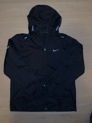  Nike windbreaker - Säljer en Nike windbreaker med huva och dragkedja framtill. Jackan har justerbar snörning i huvan och reflekterande detaljer. Perfekt för dig som vill ha en lätt och sportig jacka med stilren look.