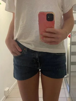 Lågmidjade jeansshorts från junkyard - Säljer ett par mörkblå denimshorts från junkyard med låg midja och klassisk femficksdesign. Shortsen har rå kant nertill och knappgylf. Tidigare jeans som är klippta till shorts. Midje mått 40 rakt över och i strl M. Perfekta för sommaren och riktigt snygga till en enkel t-shirt.