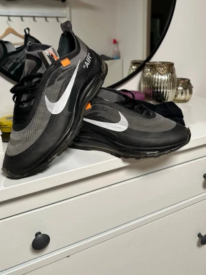 Nike Off-White Air Max 97 Svart - Säljer ett par Nike Off-White Air Max 97 i svart med vita detaljer och ikonisk Swoosh. Skorna har genomskinlig sula, textdetaljer på sidan och orange Off-White tag. Materialet är syntet med meshstruktur och snörning framtill. Riktigt snygga och unika sneakers för dig som gillar streetwear.
