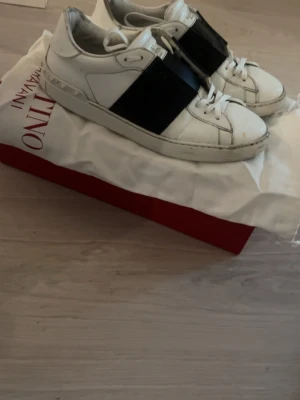 Valentino Garavani Open sneakers vit/svart - Säljer ett par Valentino Garavani Open sneakers köpta i NK i vitt skinn med bred svart läderdetalj över vristen. Klassisk rund tå, vita snören och diskret logga på plösen. Kommer med original dustbag och låda. Perfekta för dig som gillar exklusiva och stilrena sneakers.