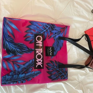 NYX Off Tropic shoppingväska rosa - Stor och färgstark shoppingväska från NYX med transparent rosa plast, svarta handtag och blå tropiska blad som mönster. Väskan har trycket 'Off Tropic' på framsidan och rymmer allt du behöver för en dag på stan eller stranden.