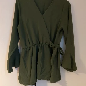 Olivgrön omlottklänning med knyt - Snygg olivgrön omlottklänning med v-ringning och knyt i midjan. Klänningen har långa ärmar med volang vid ärmslut och en lätt rynkad midja som ger en fin siluett. Perfekt för dig som gillar stilrena och trendiga plagg.