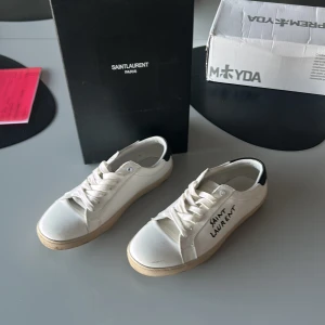 Saint Laurent vita sneakers i skinn - Snygga vita sneakers från Saint Laurent med svart detalj på hälen och handskriven logga på sidan. Klassisk låg modell med snörning och platt sula. Tillverkade i skinn och har en stilren, minimalistisk look som passar till det mesta. Kan tyvärr inte garantera äkta, köpta på Vinted för 1399