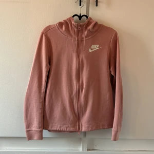 Rosa Nike hoodie med dragkedja - Säljer en snygg rosa hoodie från Nike med dragkedja framtill och huva med snörning. Tröjan har långa ärmar och vita Nike-loggor längs ryggen och ärmarna. Perfekt för chill dagar eller när du vill vara sportig.